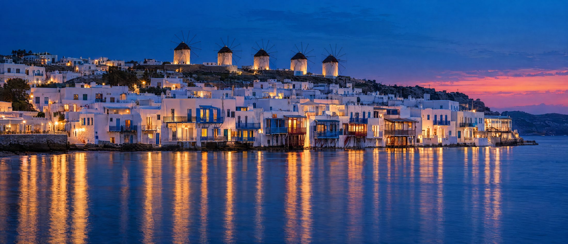 A Cycladic island town at blue hour with lit waterfront houses and windmills on the ridge